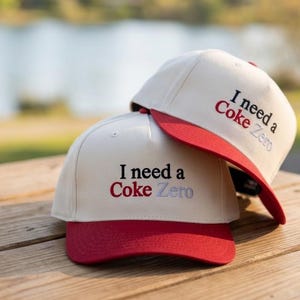 May include: Two off-white baseball caps with red brims. The front of each cap features embroidered text that reads I need a Coke Zero, with the word Coke in red and the word Zero in light blue, while the rest of the text is in black.