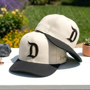 Puede incluir: Dos gorras de béisbol beige con viseras negras y una letra "D" negra en la parte delantera. Las gorras están apiladas. Las gorras están sobre una superficie blanca con un fondo borroso de vegetación y flores.