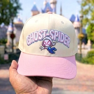 May include: A cream-colored baseball cap with a pink brim featuring an embroidered Ghost-Spider character and the text GHOST-SPIDER in purple and white lettering across the front panel. The cap has a structured crown and stitched eyelets.