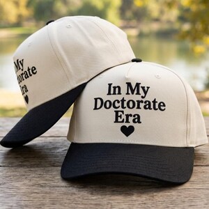 Könnte beinhalten: Zwei Baseballkappen mit cremeweißen Kronen und schwarzen Schirmen. Auf der Vorderseite der Kappen befindet sich der schwarz gestickte Text "In My Doctorate Era", gefolgt von einem kleinen schwarzen Herzsymbol. Die Kappen haben ein strukturiertes Design.