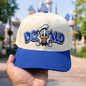 Könnte beinhalten: Eine cremefarbene Baseballkappe mit blauem Schirm und einer blauen, gestickten Donald Duck-Grafik auf der Vorderseite. Der Schriftzug "DONALD" ist in blauen Buchstaben um die Figur herum gestickt und zeigt ein klassisches Cartoon-Design.