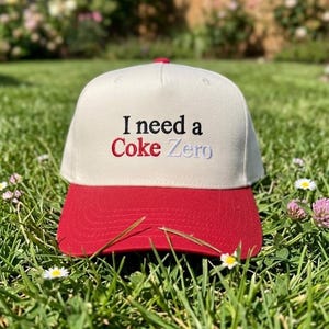 May include: A cream-colored baseball cap with a red brim. The front of the cap features embroidered text that reads "I need a" in black, "Coke" in red, and "Zero" in light blue, centered on the crown above the red visor.