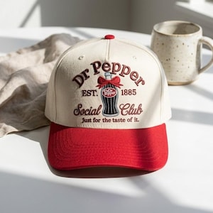 May include: A cream-colored baseball cap with a red brim featuring embroidered text that reads Dr Pepper EST. 1885 Social Club Just for the taste of it. The design includes a graphic of a soda bottle with a red bow tied around the neck.