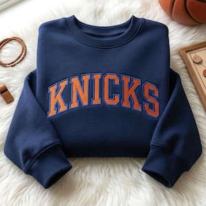 May include: A navy blue crewneck sweatshirt featuring the word KNICKS in large, arched orange letters with a blue outline. The garment has long sleeves with ribbed cuffs and a ribbed neckline, showcasing a classic athletic design.