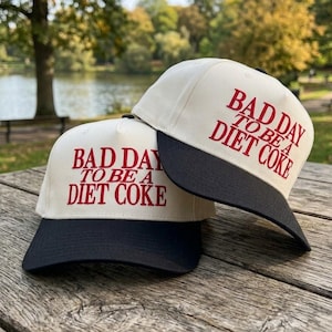 May include: Two baseball caps with a cream-colored crown and a black brim. The caps have the text "BAD DAY TO BE A DIET COKE" in red. The caps are on a wooden picnic table.