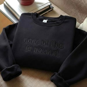 May include: A black long-sleeve crewneck sweatshirt featuring the embroidered text "none of this is normal" in a matching black thread across the chest. The garment has a ribbed collar, cuffs, and hem, with a visible white interior brand label.