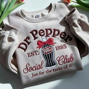 Pode incluir: Camisola bege com o logótipo Dr Pepper bordado em bordô e preto. O design inclui uma garrafa com um laço vermelho, o texto "Dr Pepper", "EST. 1885" e "Social Club". A frase "Just for the taste of it" também está incluída.