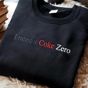 Puede incluir: Sudadera negra de cuello redondo con la frase bordada "I need a Coke Zero". El texto está en una combinación de hilo gris, rojo y blanco. La sudadera está doblada y colocada sobre una superficie suave y clara.
