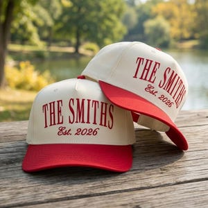 Op de afbeelding: Twee crèmekleurige baseballpetten met rode klepjes liggen op een houten oppervlak. De petten hebben de tekst "THE SMITHS" in rood schrift, met "Est. 2026" eronder. De achtergrond toont een wazig uitzicht op een meer en bomen.
