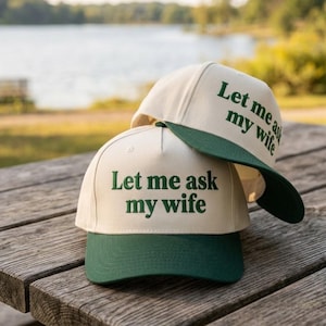 Op de afbeelding: Twee baseballpetten met een gebroken witte kroon en een donkergroene klep. Op de voorkant van elke pet staat de geborduurde tekst Let me ask my wife in donkergroene letters. De petten zijn gestructureerd en hebben een knoop aan de bovenkant.
