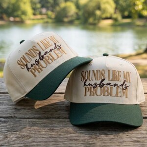 May include: Two baseball caps with cream-colored crowns and dark green brims. The front of each cap features embroidered text in gold and dark brown that reads, "SOUNDS LIKE MY husband's PROBLEM," with the word "husband's" in a cursive script.