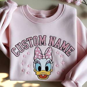 Peut inclure: Un sweat-shirt rose clair avec le texte "CUSTOM NAME" en arc de cercle. En dessous, un dessin animé de canard avec un nœud rose à pois blancs. Des étoiles roses brodées entourent le canard.