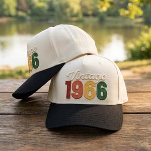May include: Two baseball caps with off-white crowns and black brims. The front of each cap features embroidered text that reads Vintage in a script font above the number 1966, with each digit stitched in a different color: red, orange, yellow, and green.