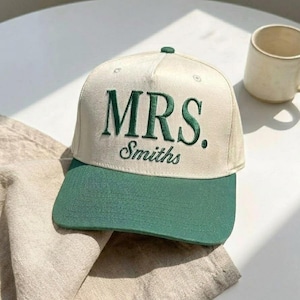 May include: A cream-colored baseball cap with a green brim. The cap features the embroidered text "MRS. Smiths" in green. A small green button is on top of the cap. A beige mug is in the background.