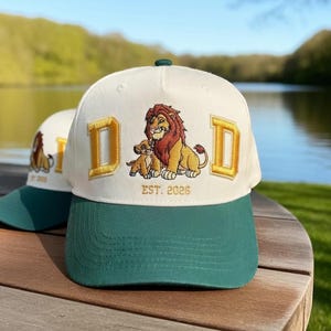 Puede incluir: Gorra de béisbol blanca con visera verde y letras "D" doradas. La gorra presenta un diseño bordado de un león y su cachorro con el texto "EST. 2026". La gorra está sobre una superficie de madera.