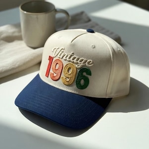 Può includere: Cappellino da baseball color crema con visiera blu navy. Il cappellino presenta la scritta ricamata "Vintage 1996" in rosso, giallo e verde. Un piccolo bottone blu sulla parte superiore.