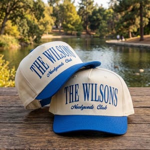 Pode incluir: Dois bonés de beisebol com copa creme e aba azul. Os bonés têm a inscrição "THE WILSONS Newlyweds Club" bordada em azul. Os bonés estão sobre uma superfície de madeira.