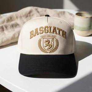 May include: Cream and black baseball cap with gold embroidered text reading "BASGIATH WAR COLLEGE" and a dragon crest. A classic cap design, suitable for casual wear. The cap is a stylish accessory.
