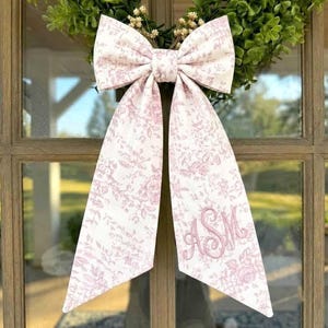 May include: A large fabric bow with a light pink and white floral toile pattern, featuring two long, pointed tails. The right tail displays an embroidered monogram in pink script. The bow is attached to a green leafy wreath on a glass door.