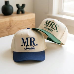 Puede incluir: Dos gorras de béisbol con coronas de color crema. Una gorra presenta un bordado azul oscuro que dice MR. Smiths con una visera a juego. La otra gorra presenta un bordado verde oscuro que dice MRS. Smiths con una visera a juego.
