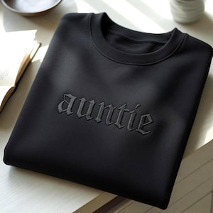Pode incluir: Uma camisola preta de gola redonda com a palavra auntie bordada em fio preto a condizer no peito. O texto é apresentado num tipo de letra gótico arrojado, criando um aspeto subtil e monocromático na superfície lisa do tecido.