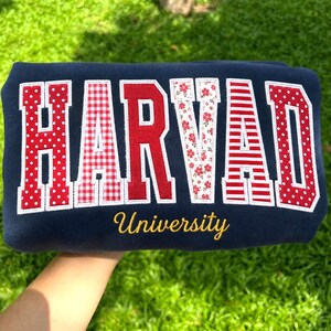 Könnte beinhalten: Ein marineblaues Sweatshirt mit dem Wort HARVAD in großen Blockbuchstaben. Jeder Buchstabe hat ein einzigartiges rot-weißes Muster, darunter Punkte, Vichy-Karo, einfarbiges Rot, Blumen und Streifen. Das Wort University ist in Gelb gestickt.