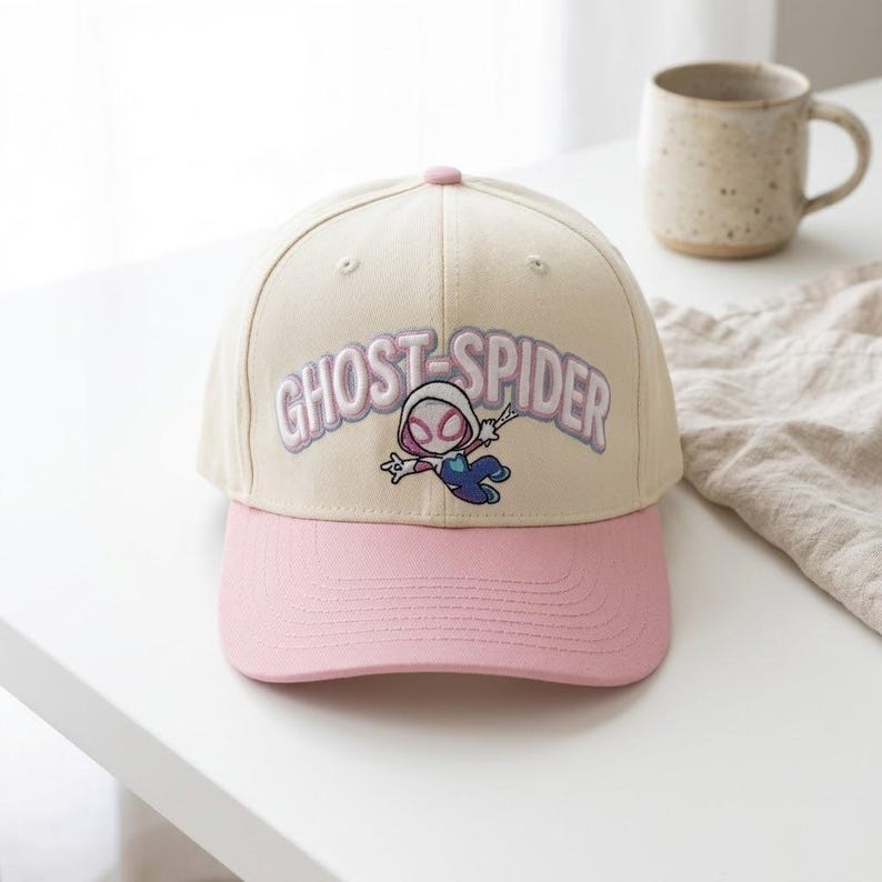 May include: A cream-colored baseball cap with a pink brim. The front features an embroidered graphic of a cartoon character in a white, pink, and blue suit, with the text GHOST-SPIDER arched above in light purple lettering with a white outline.