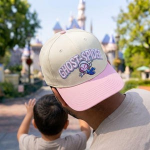 May include: A beige baseball cap with a pink brim featuring an embroidered Ghost-Spider character and the text GHOST-SPIDER in purple lettering. The cap has a button on top and stitched eyelets on the crown, showcasing a structured design.