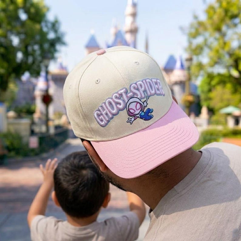 May include: A beige baseball cap with a pink brim featuring an embroidered Ghost-Spider character and the text GHOST-SPIDER in purple lettering. The cap has a button on top and stitched eyelets on the crown, showcasing a structured design.