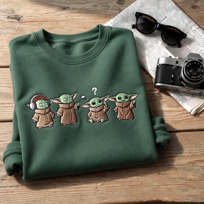 May include: Green sweatshirt featuring an embroidered design of four Grogu characters in various poses. The characters are wearing brown robes and have large ears. One character has headphones. A question mark is above one character's head.