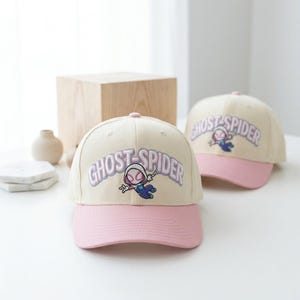 May include: Two baseball caps with cream-colored crowns and pink brims. Each cap features an embroidered graphic of the character Ghost-Spider in a dynamic pose, accompanied by the text GHOST-SPIDER in large, light purple, arched lettering.