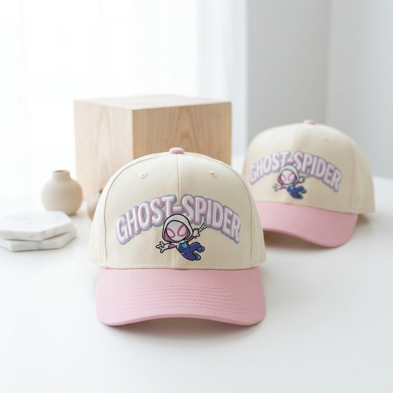 May include: Two baseball caps with cream-colored crowns and pink brims. Each cap features an embroidered graphic of the character Ghost-Spider in a dynamic pose, accompanied by the text GHOST-SPIDER in large, light purple, arched lettering.