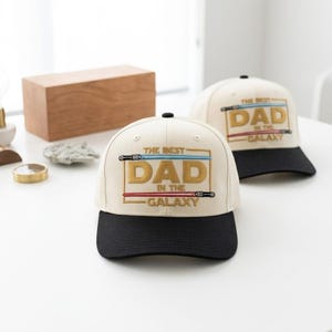 May include: Two baseball caps with the text "THE BEST DAD IN THE GALAXY" embroidered in gold, blue, and red. The caps have a cream-colored crown and a black brim. The caps are a fun gift idea.