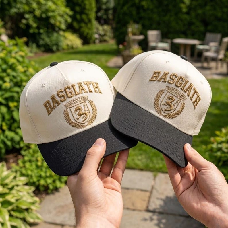 May include: Two cream baseball caps with black peaks. Each cap displays the embroidered text "BASGIATH War College" above a crest with a lion. The caps are held up against a green, leafy background.