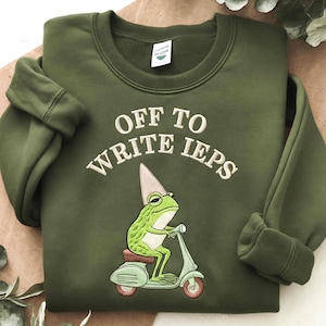 以下が含まれることがあります： オリーブグリーンのクルーネックトレーナー。白い刺繍で「OFF TO WRITE IEPS」と書かれています。その下には、ピンクのパーティーハットをかぶり、茶色の車輪がついたライトグリーンのスクーターに乗った緑色のカエルの刺繍が施されています。