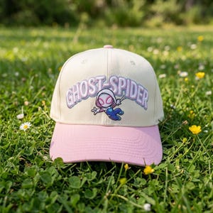 May include: A cream-colored baseball cap with a pink brim featuring an embroidered Ghost-Spider character graphic and the text GHOST-SPIDER in light purple lettering with a white outline centered on the front panel of the hat.