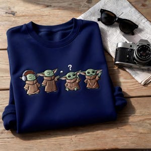 May include: Navy blue sweatshirt featuring an embroidered design of four Grogu characters in various poses. The design includes a Grogu with headphones, one with a ball, one with a question mark, and one waving. Sunglasses and a vintage camera are also visible.
