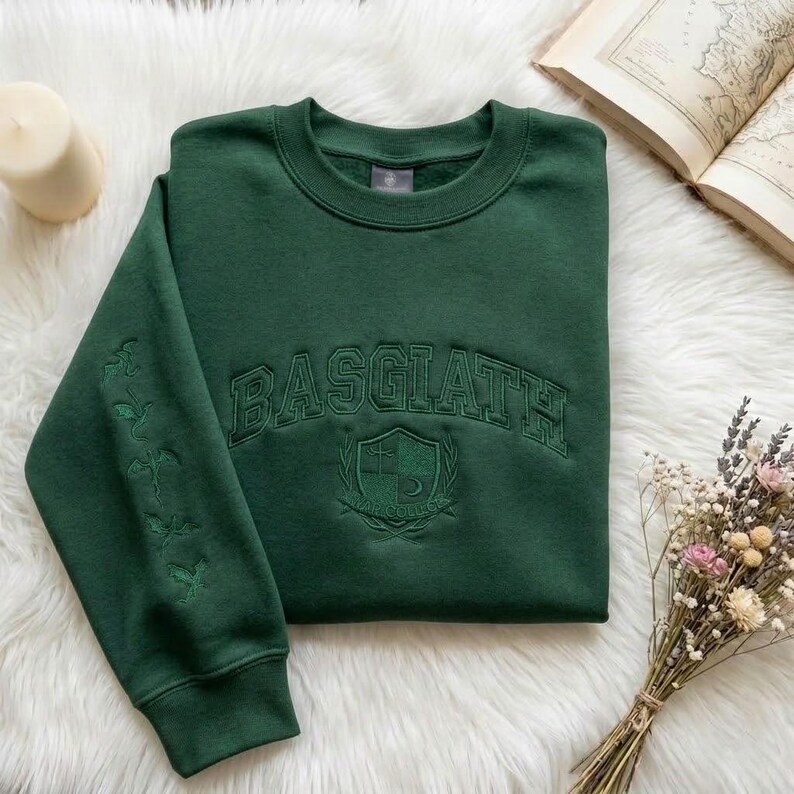 May include: A dark green sweatshirt with the embroidered word "BASGIATH" across the chest. Below the word is a crest design. The left sleeve has embroidered floral designs. The sweatshirt is on a white fluffy surface.