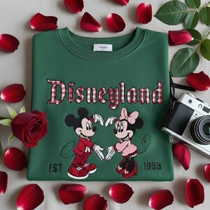 May include: Green sweatshirt with the word "Disneyland" in red plaid lettering. Features embroidered images of Mickey and Minnie Mouse. The sweatshirt is surrounded by red rose petals and a vintage camera.