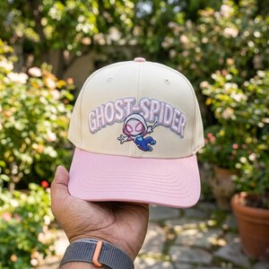 May include: A cream-colored baseball cap with a pink brim featuring an embroidered Ghost-Spider character graphic and the text GHOST-SPIDER in light purple lettering across the front panel, designed with a structured crown and top button.