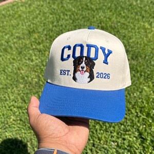 May include: A cream-colored baseball cap with a bright blue brim and a blue button on top. The front features embroidered blue text that reads CODY, EST. 2026, and a detailed embroidered portrait of a Bernese Mountain Dog with its tongue out.