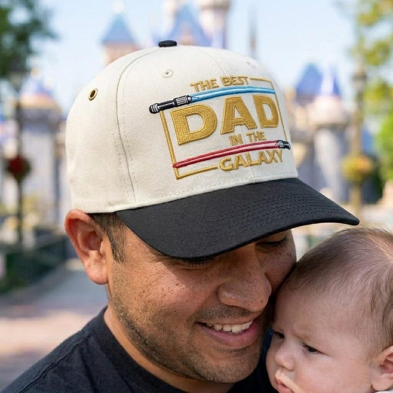 May include: Cream-colored baseball cap with a black brim. The cap features the text "THE BEST DAD IN THE GALAXY" in gold and red embroidery, with lightsaber designs. A stylish accessory for any dad.