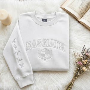 May include: A white sweatshirt with the word "BASGIATH" arched across the chest. Below the text is a crest design. The left sleeve features embroidered dragon silhouettes. The sweatshirt is folded on a white, fluffy surface.