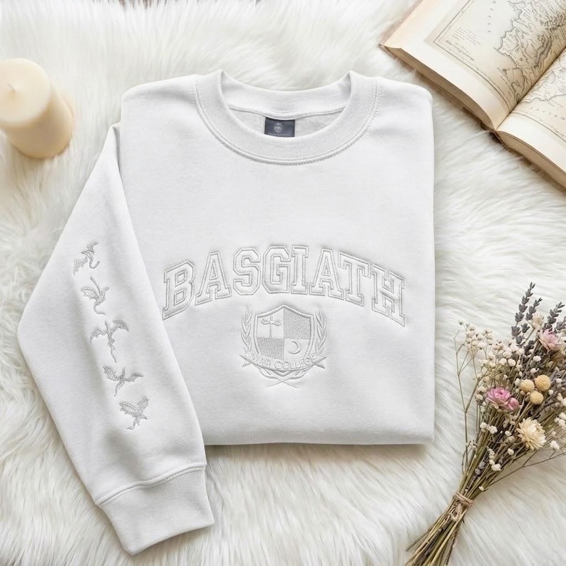 May include: A white sweatshirt with the word "BASGIATH" arched across the chest. Below the text is a crest design. The left sleeve features embroidered dragon silhouettes. The sweatshirt is folded on a white, fluffy surface.