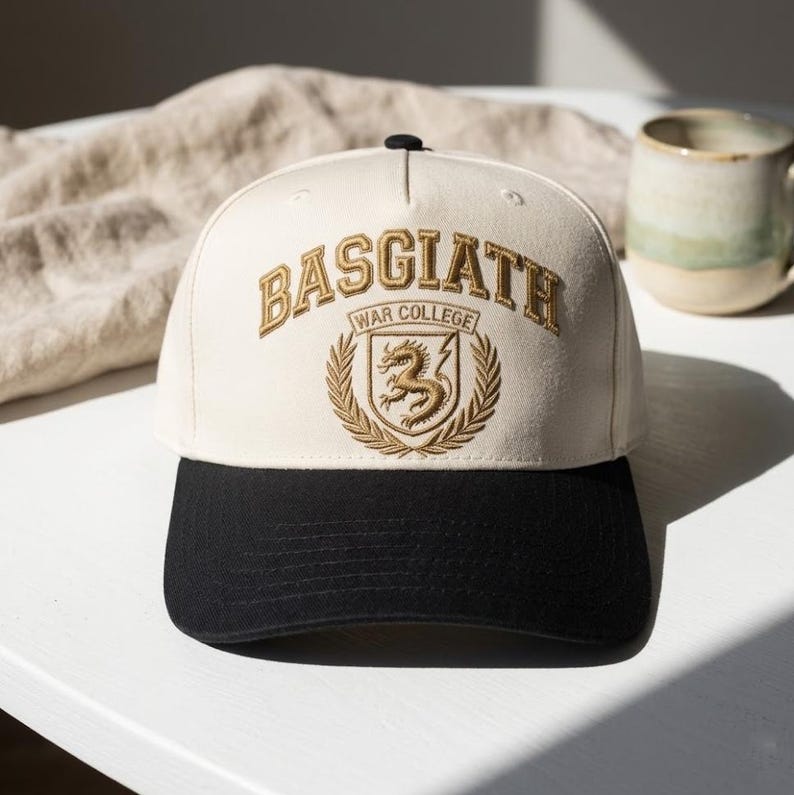 May include: Cream and black baseball cap with gold embroidered text reading "BASGIATH WAR COLLEGE" and a dragon crest. A classic cap design, suitable for casual wear. The cap is a stylish accessory.