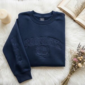 May include: A navy blue sweatshirt featuring the embroidered word "BASGIATH" across the chest, with a crest below and bird designs on the left sleeve. The sweatshirt is displayed on a white, fluffy surface. The text "WAR COLLEGE" is below the crest.
