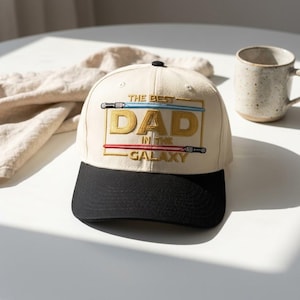 May include: Cream-colored baseball cap with a black brim. The cap features the text "THE BEST DAD IN THE GALAXY" in gold and white, with blue and red lightsaber designs. A stylish accessory for any dad.