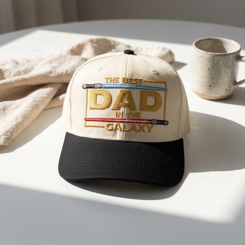 May include: Cream-colored baseball cap with a black brim. The cap features the text "THE BEST DAD IN THE GALAXY" in gold and white, with blue and red lightsaber designs. A stylish accessory for any dad.