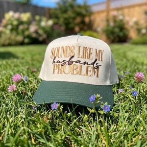May include: A baseball cap with a cream-colored crown and a dark green brim. The front of the crown features embroidered text in gold and dark brown that reads, "SOUNDS LIKE MY husbands PROBLEM" in a mix of block and cursive lettering.