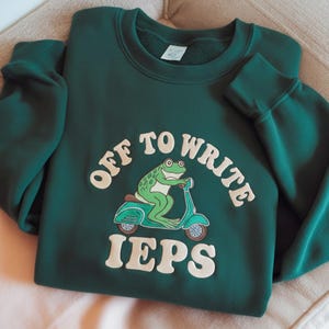 Peut inclure: Un sweat-shirt vert foncé avec l'inscription blanche "OFF TO WRITE IEPS", et une grenouille de dessin animé sur un scooter turquoise. La grenouille est verte avec un siège marron. Le sweat-shirt est plié sur une surface claire.